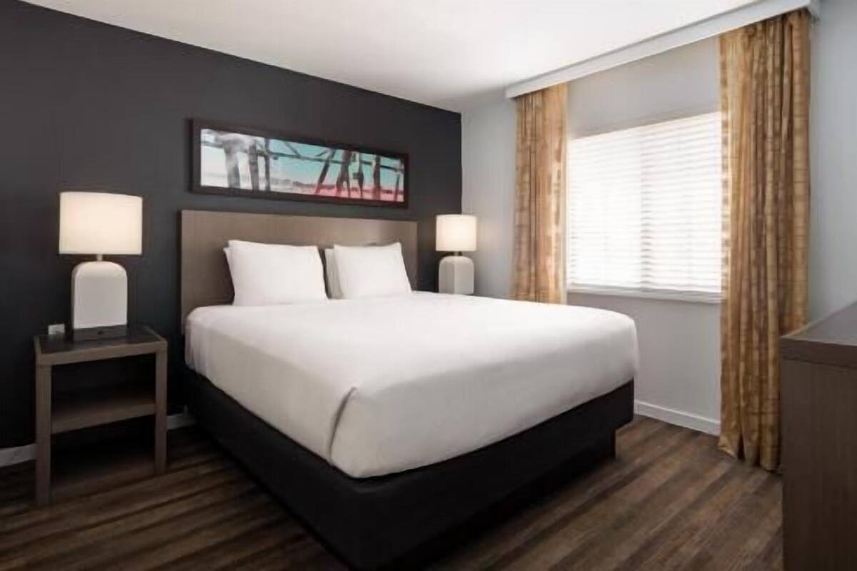 A hotel room with a bed, two bedside lamps, a painting above the bed, and a window with curtains.