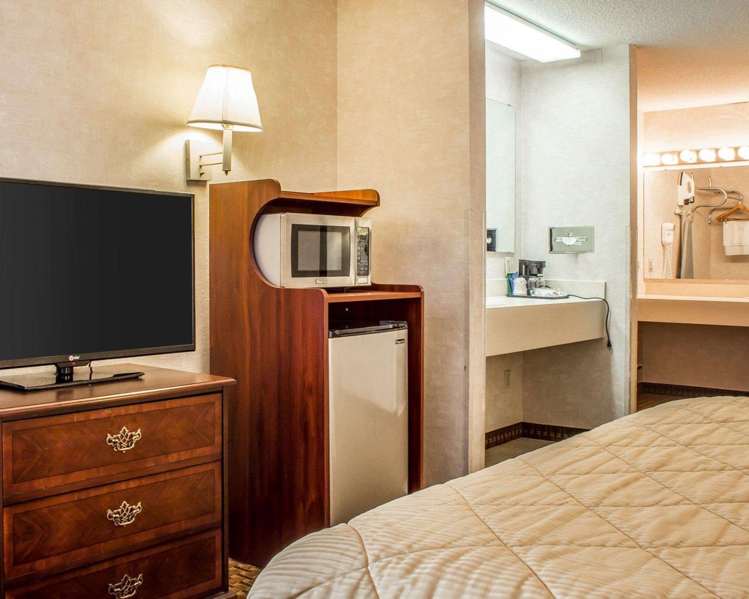 A hotel room with a bed, a TV, a microwave, a mini-fridge, a vanity with a mirror, and a lamp.