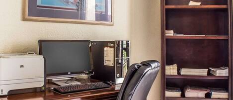 A wooden desk with a desktop computer, a printer, and a leather chair. A bookshelf with books and papers is to the right.