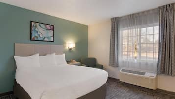 Habitación de hotel con una cama grande, una mesita de noche con lámpara, una silla, una ventana con cortinas y un aire acondicionado instalado en la pared.