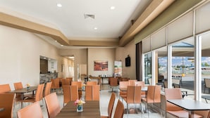 A spacious dining area with multiple tables and chairs, a kitchen area in the background, and large windows offering a view of the outside.