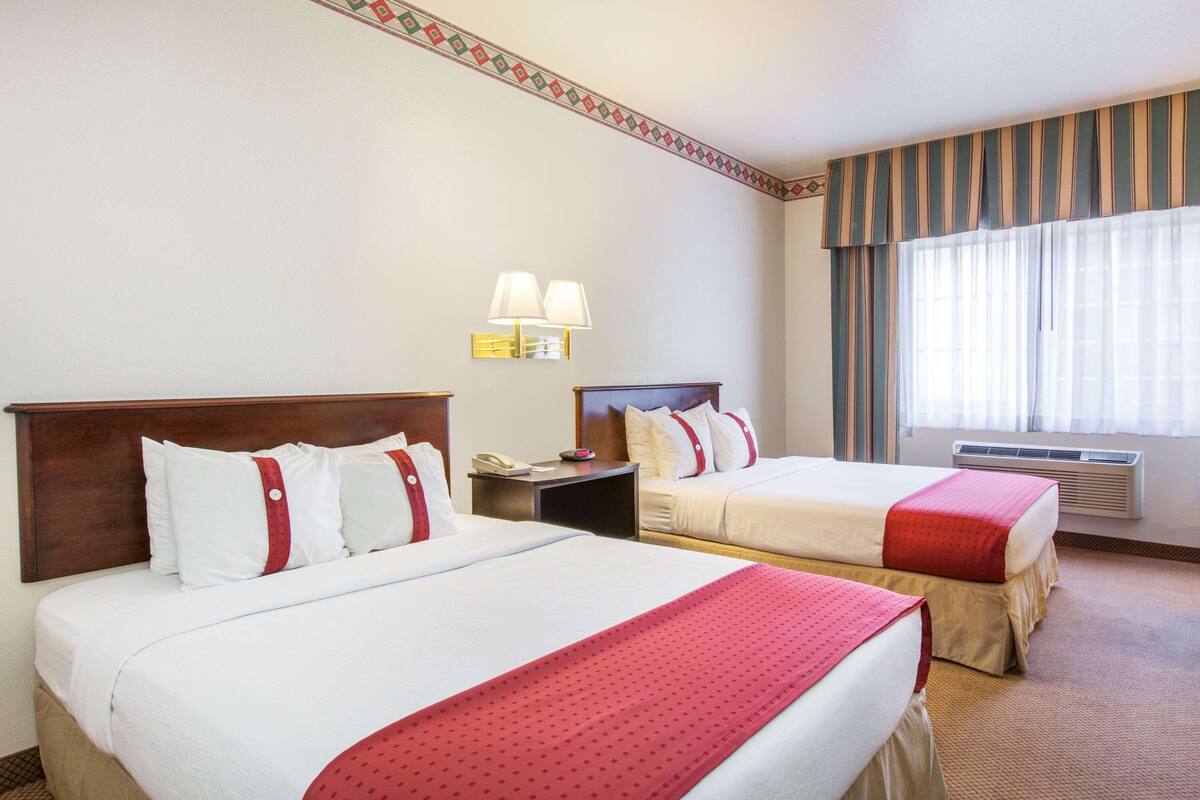A hotel room with two beds, a nightstand, a lamp, and a window with curtains.