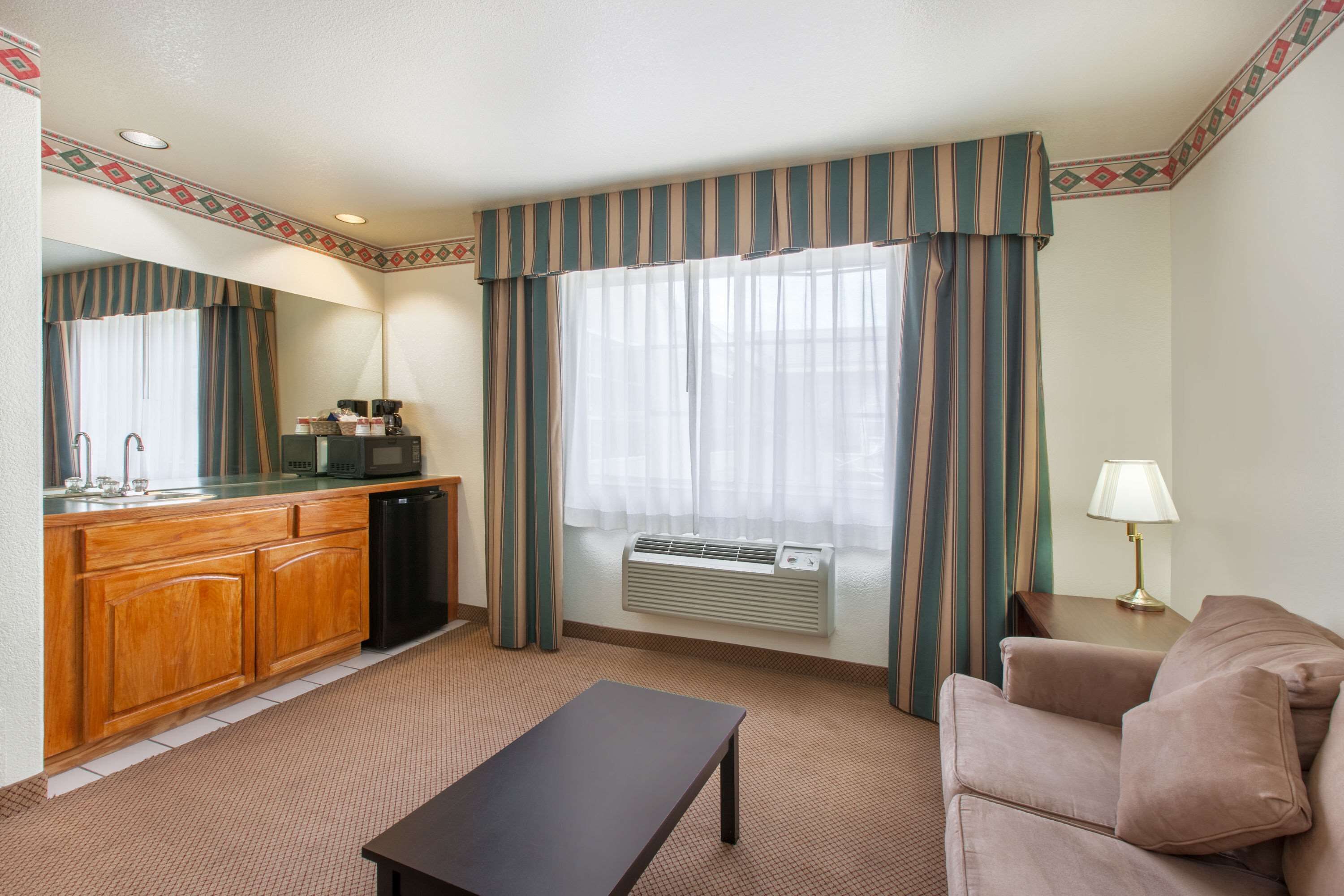 A hotel room with a kitchenette, a sofa, a table, and a large window with curtains.