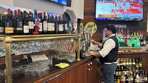 A bar area with a variety of liquor bottles and glasses, a television displaying a sports game, and a bar counter with a bartender. - Ramada by Wyndham Williams/Grand Canyon Area (Williams)