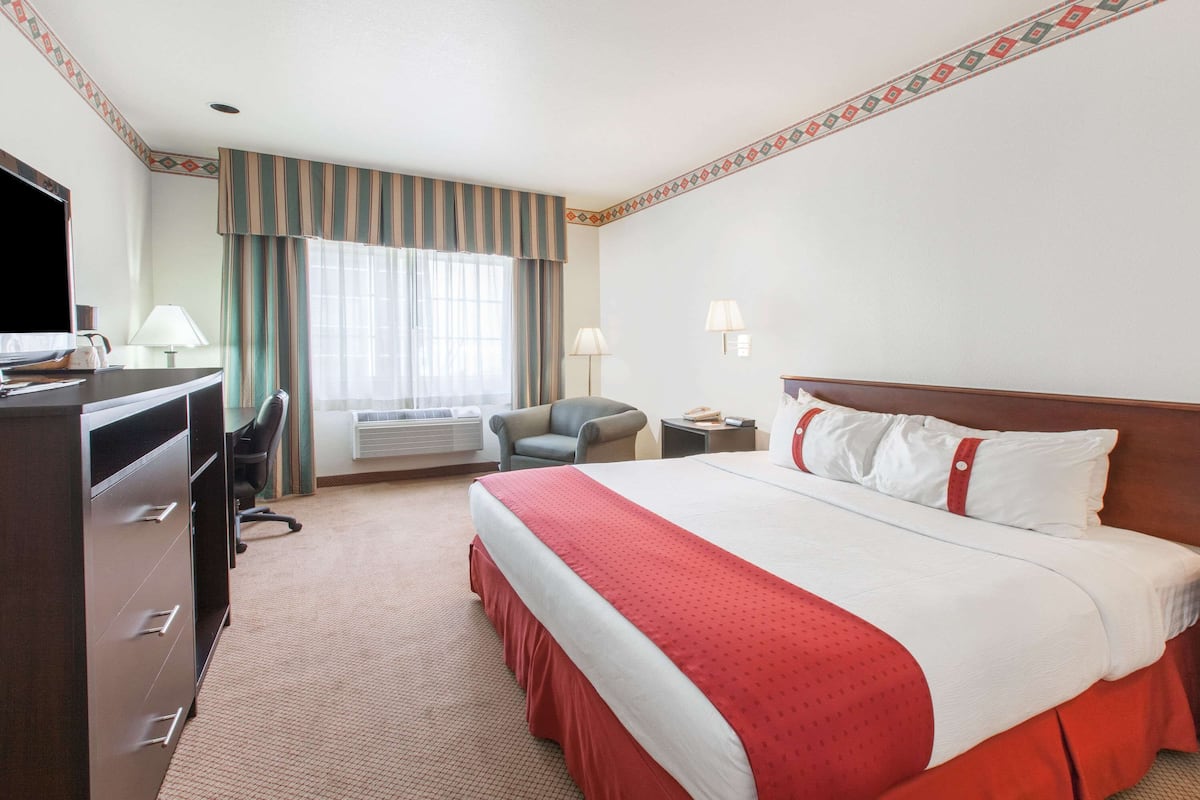 A hotel room with a large bed, a desk with a computer, a chair, a lamp, and a window with curtains.