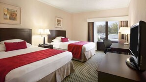 A hotel room with two beds, a desk with a TV, and a window with a view of parked cars. - Ramada by Wyndham Williams/Grand Canyon Area (Williams)