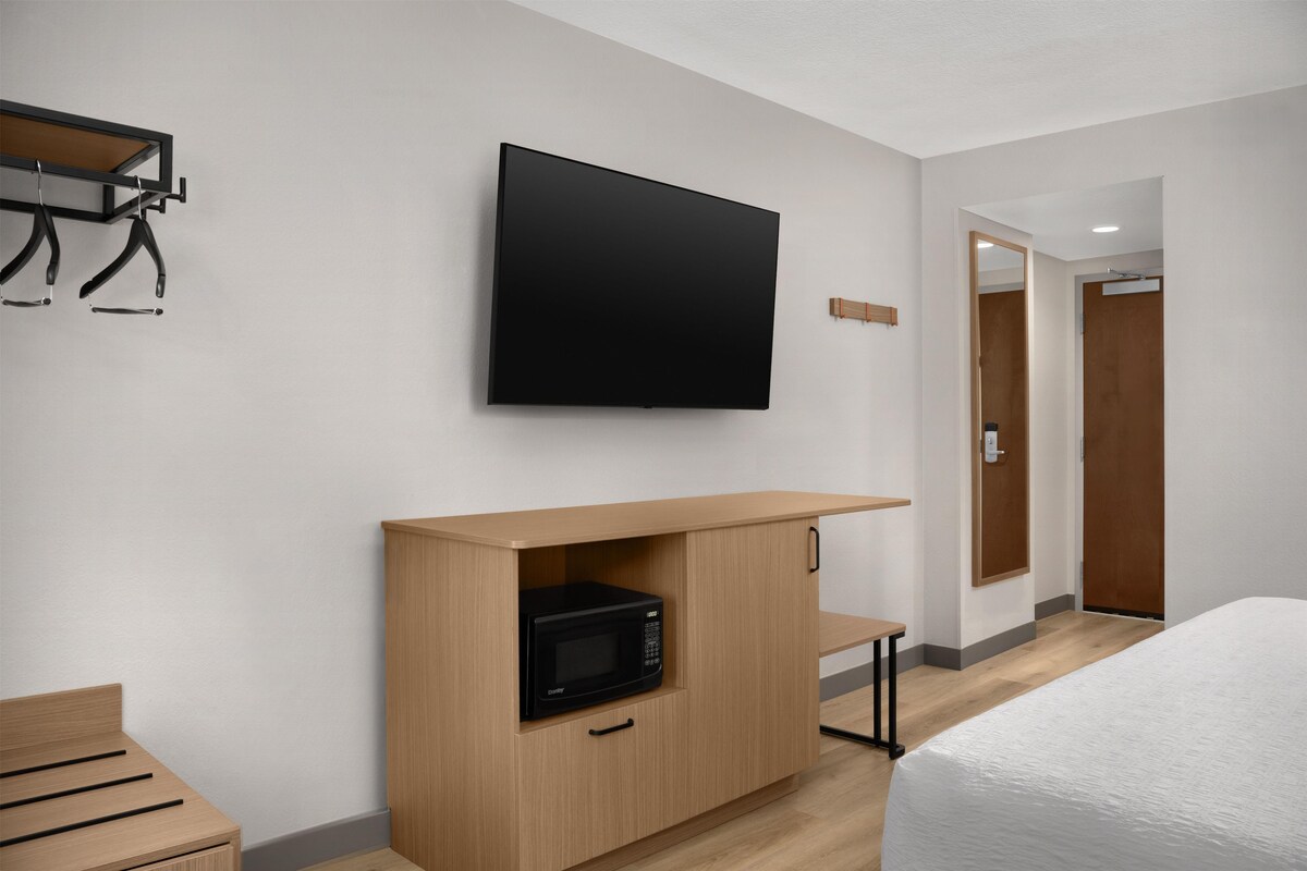 A hotel room with a flat-screen TV mounted on the wall, a wooden cabinet with a microwave, a bed, and a small table.