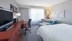 Una habitación de hotel con cama, escritorio con televisor, silla, sofá y ventana con cortinas.