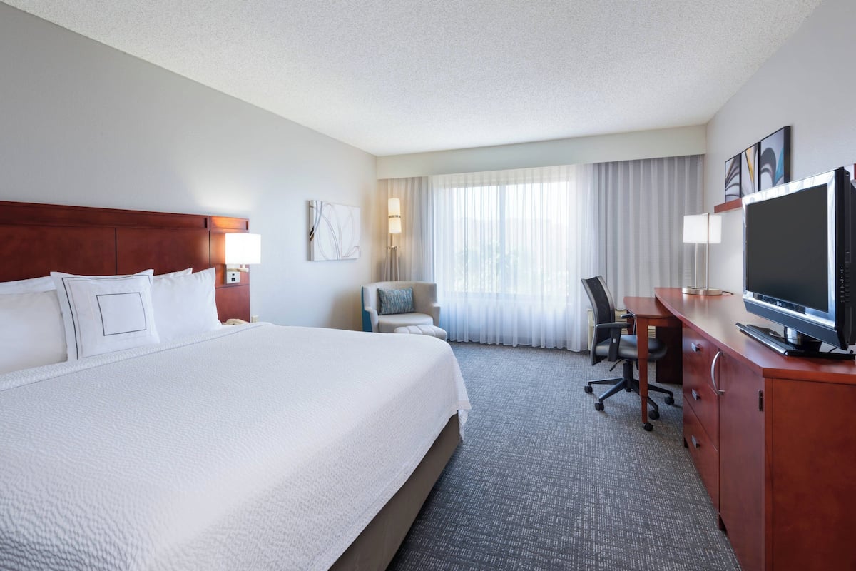 A hotel room with a large bed, a desk with a television, a chair, and a lamp.