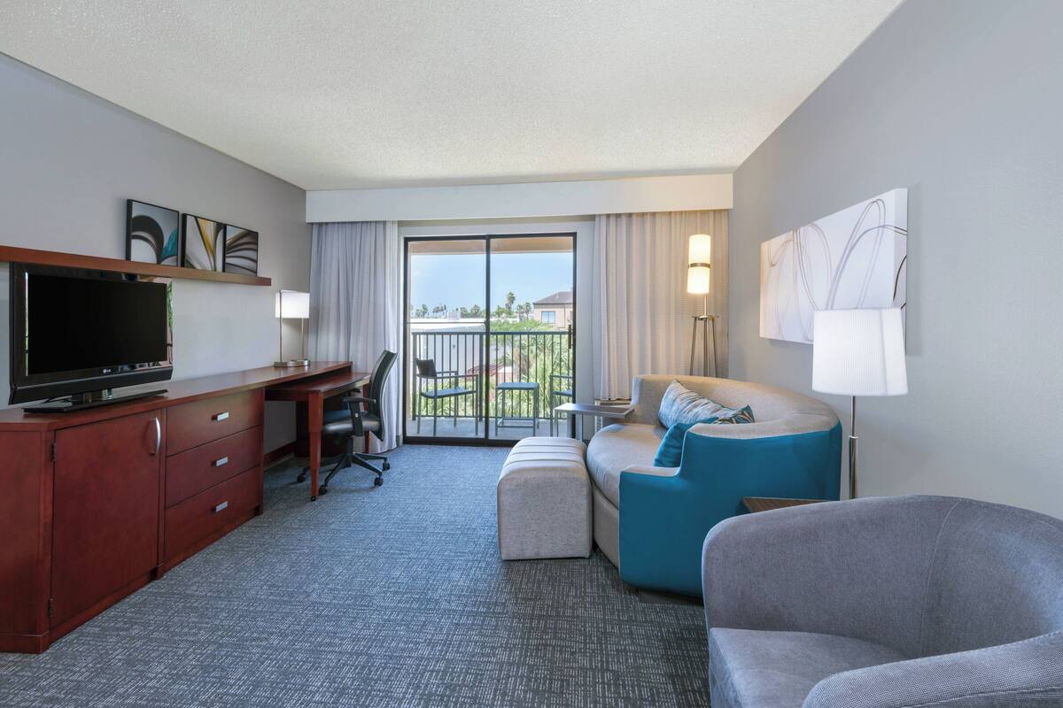 A hotel room with a desk, television, sofa, and balcony.