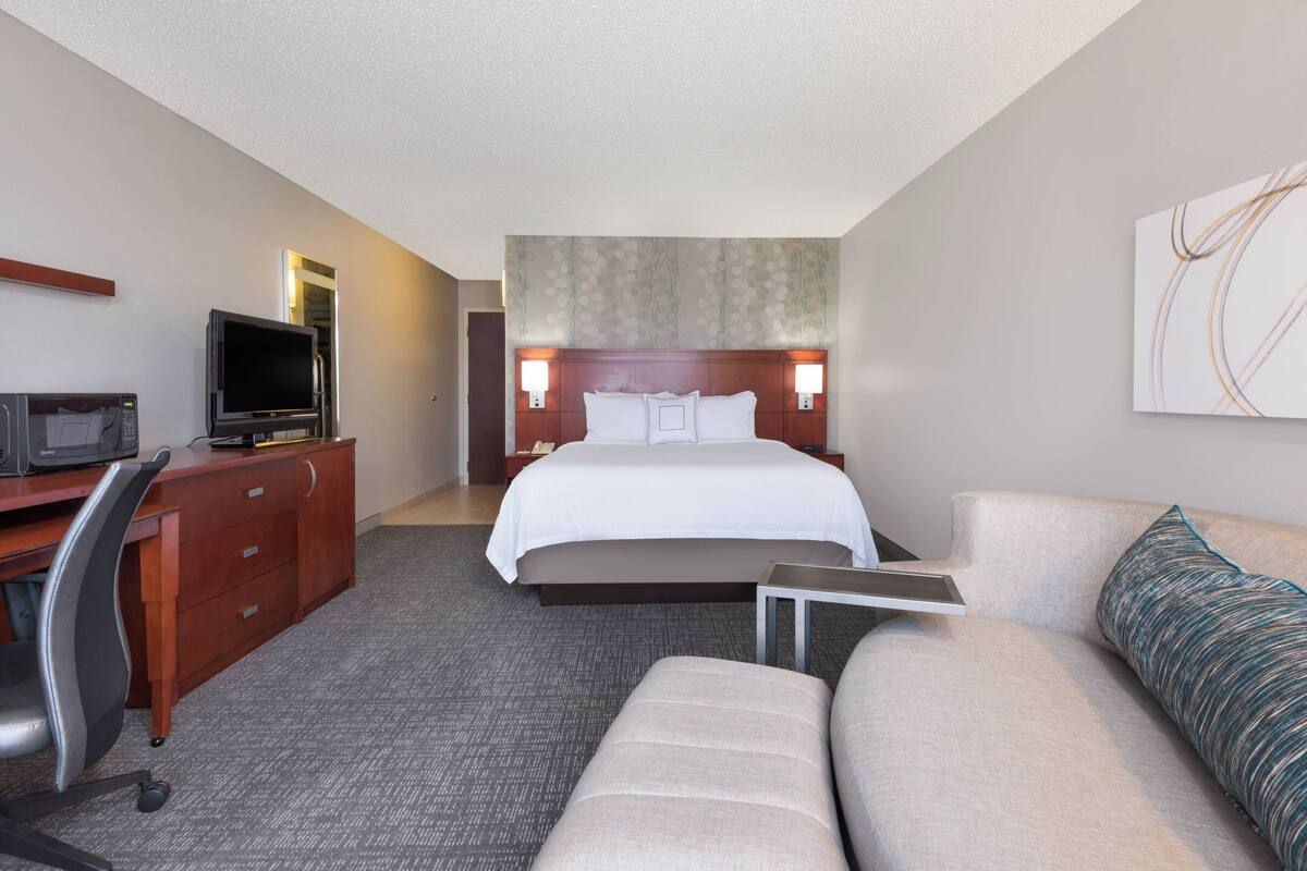 A hotel room with a bed, a desk with a television, a sofa, and a headboard with wall sconces.