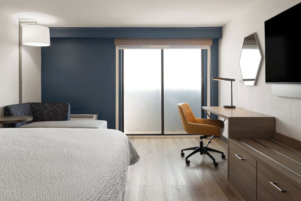A hotel room with a large bed, a desk, a chair, a TV, and a sliding door to the outside.