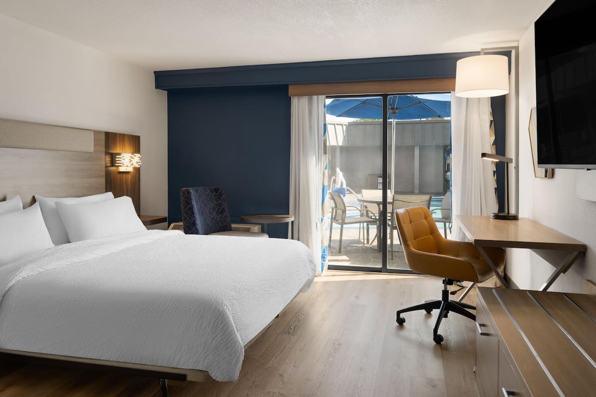 A hotel room with a bed, desk, chair, and a view through a glass door.