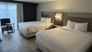 Habitación de hotel con dos camas individuales, una mesita pequeña, una silla y un ventanal grande con cortinas.