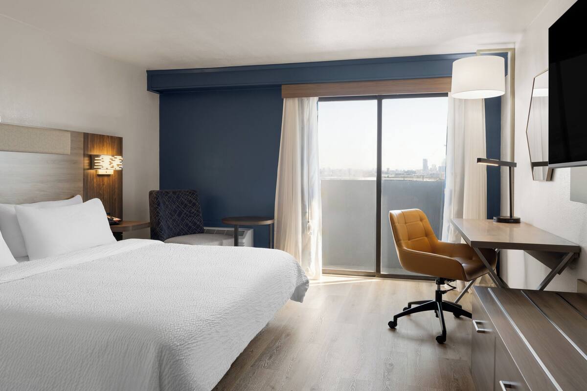 A hotel room with a large bed, a desk, a chair, a TV, and a view of the city through the window.