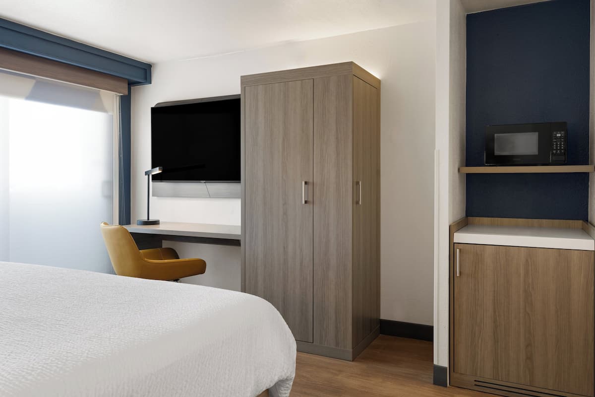 A hotel room with a bed, a desk with a chair, a wardrobe, and a microwave on a shelf.
