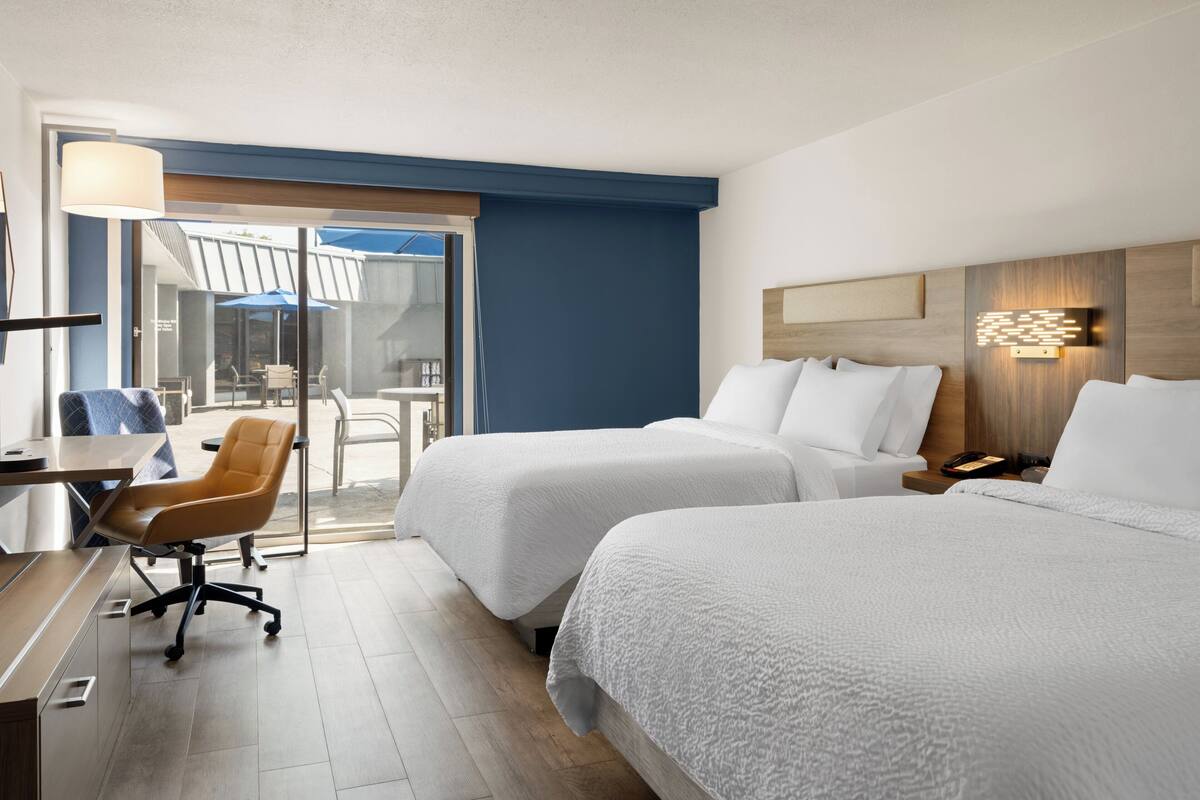 A hotel room with two beds, a desk, a chair, and a view of the outdoors.