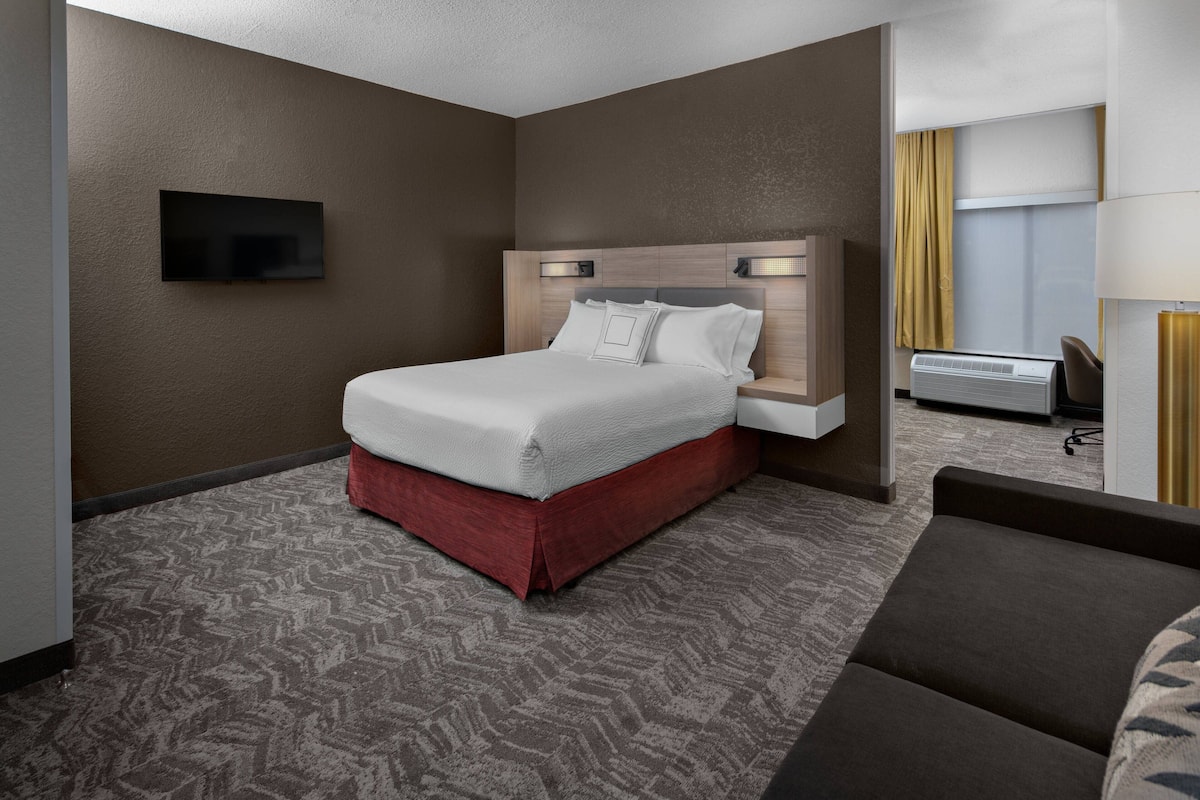 A hotel room with a large bed, a flat-screen TV mounted on the wall, a desk with a chair, and an air conditioning unit.
