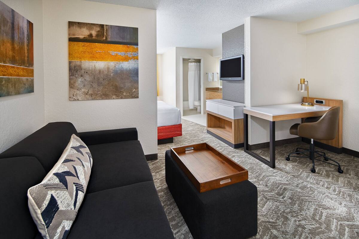 A modern hotel room with a black sofa, a wooden side table, a desk with a chair, a TV mounted on the wall, and artwork on the walls.