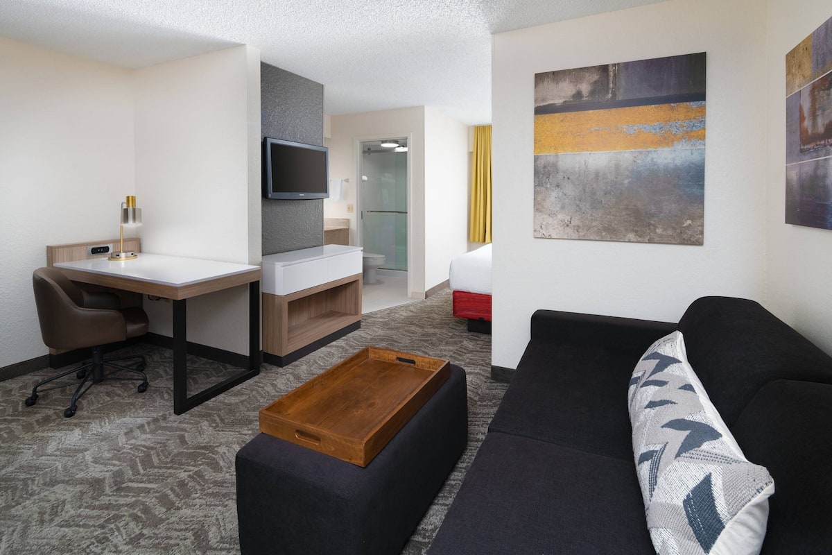 A modern hotel room with a desk, a sofa, a TV mounted on the wall, and a painting.