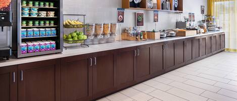 Espaço de café da manhã de hotel, com máquina de bebidas automática, potes de frutas e diversas opções de comida em um buffet.