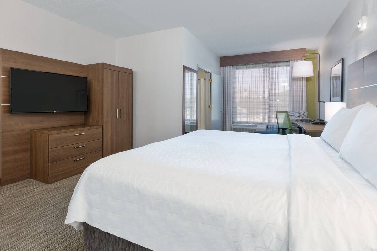A hotel room with a large bed, a flat-screen TV mounted on a wooden cabinet, a desk with a chair, and a window with curtains.