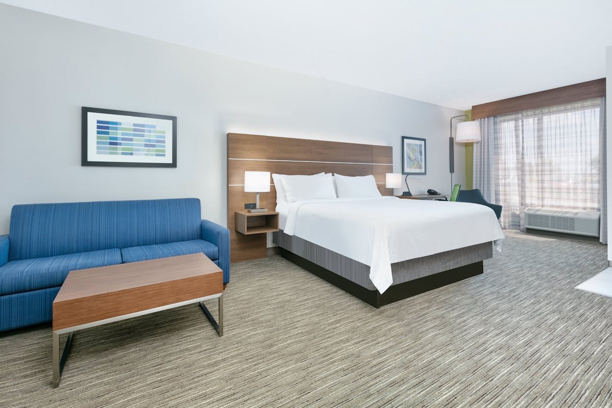A hotel room with a bed, a blue sofa, a wooden coffee table, a nightstand with a lamp, and a window with curtains.