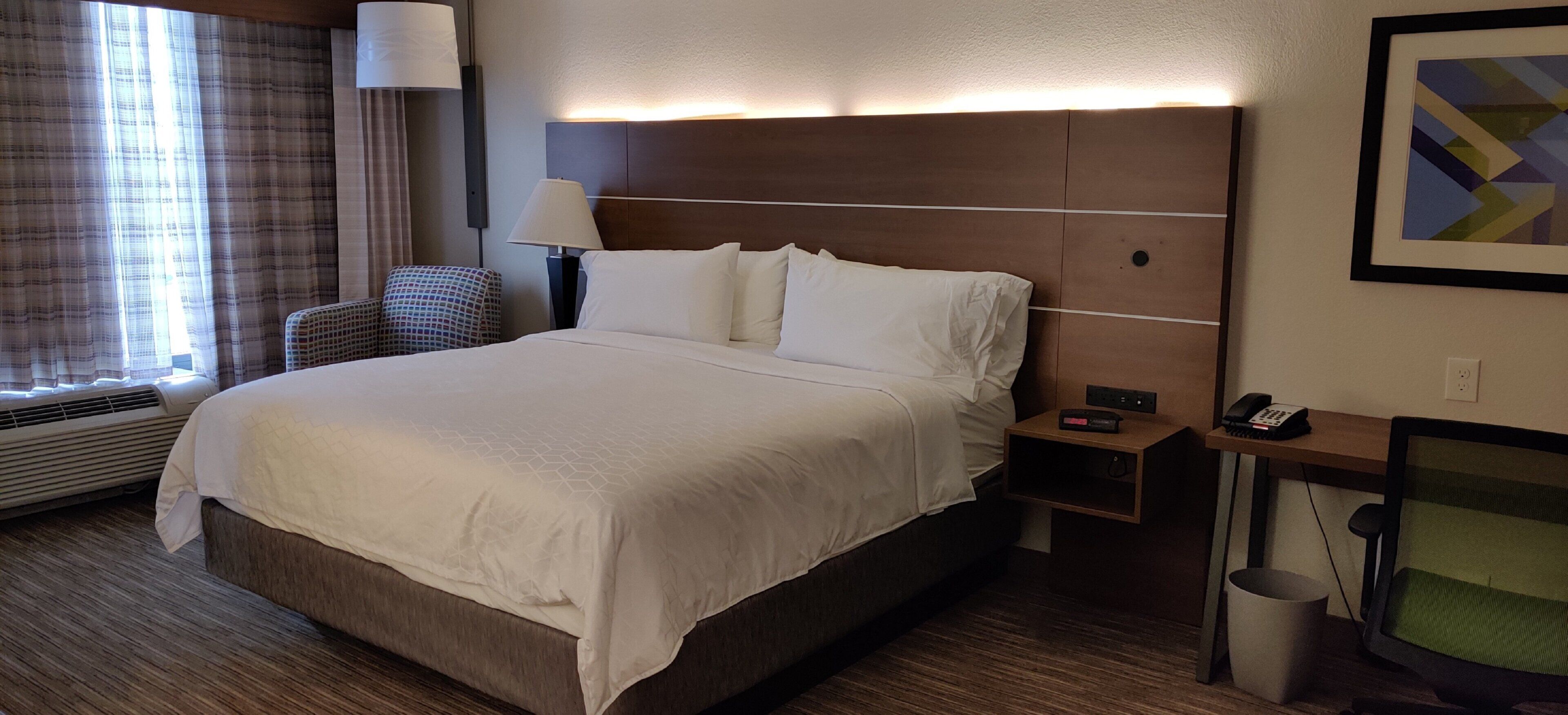 A hotel room with a large bed, a bedside table with a lamp, a chair, and a wall-mounted phone.