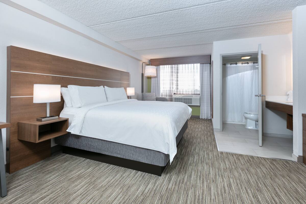 A modern hotel room with a large bed, bedside tables, a bathroom with a shower, and a view of the outdoors.