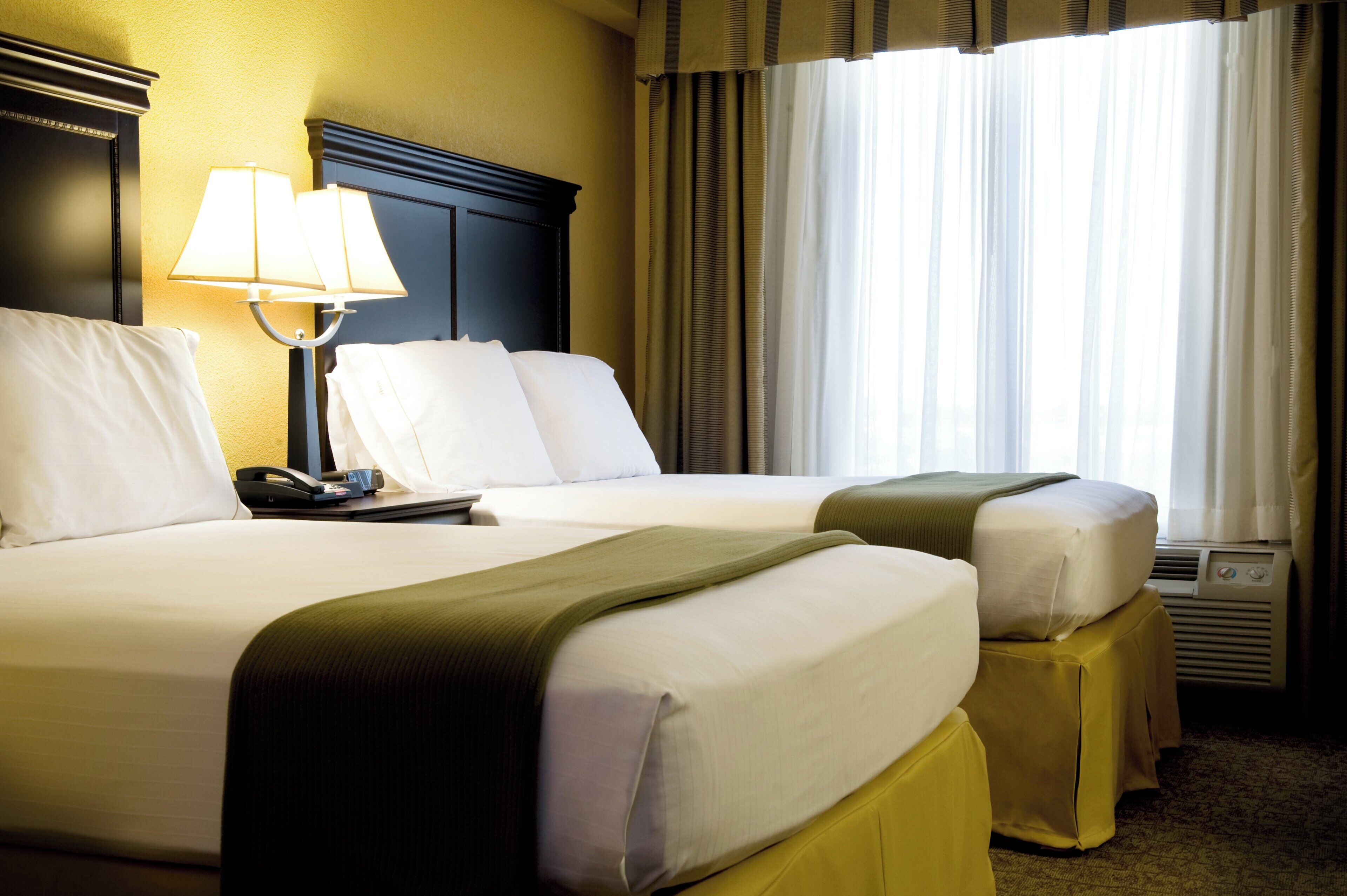A hotel room with two beds, a bedside table with a lamp, a telephone, and a window with curtains.