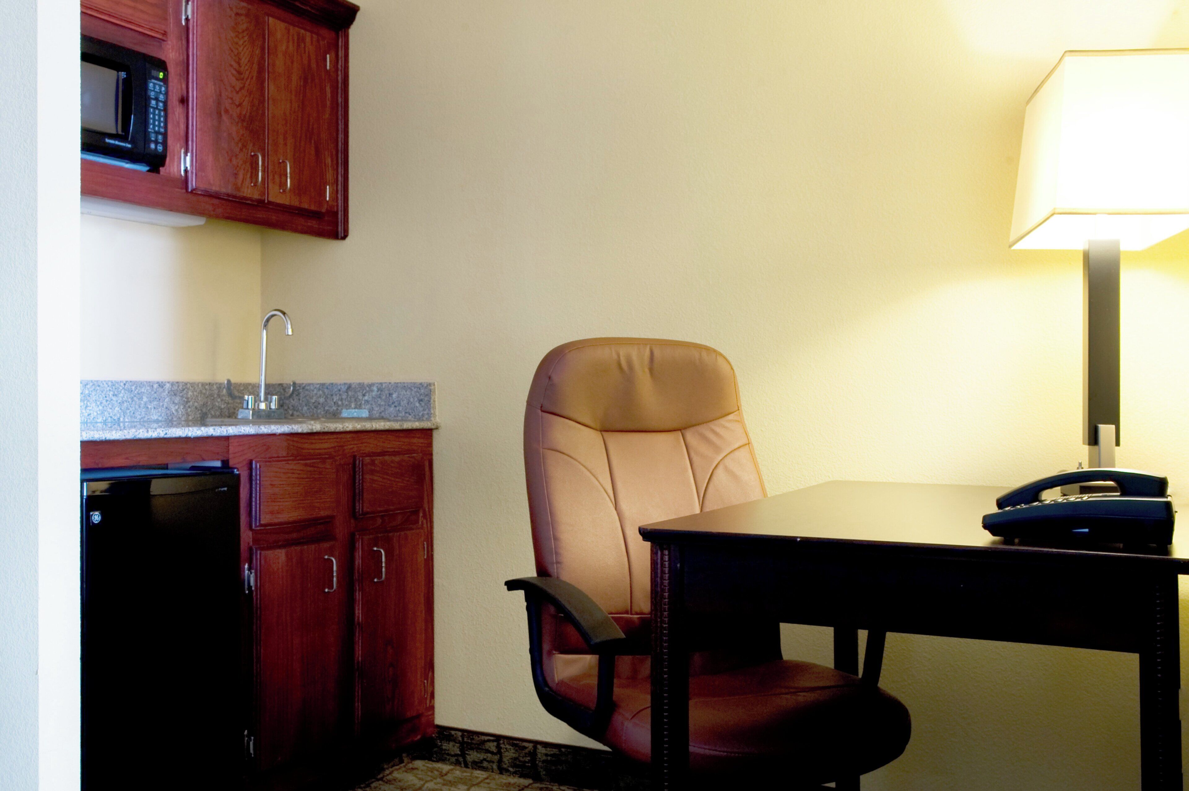 A small kitchenette with a microwave, sink, and cabinets. There is a chair and a table with a lamp and a telephone.