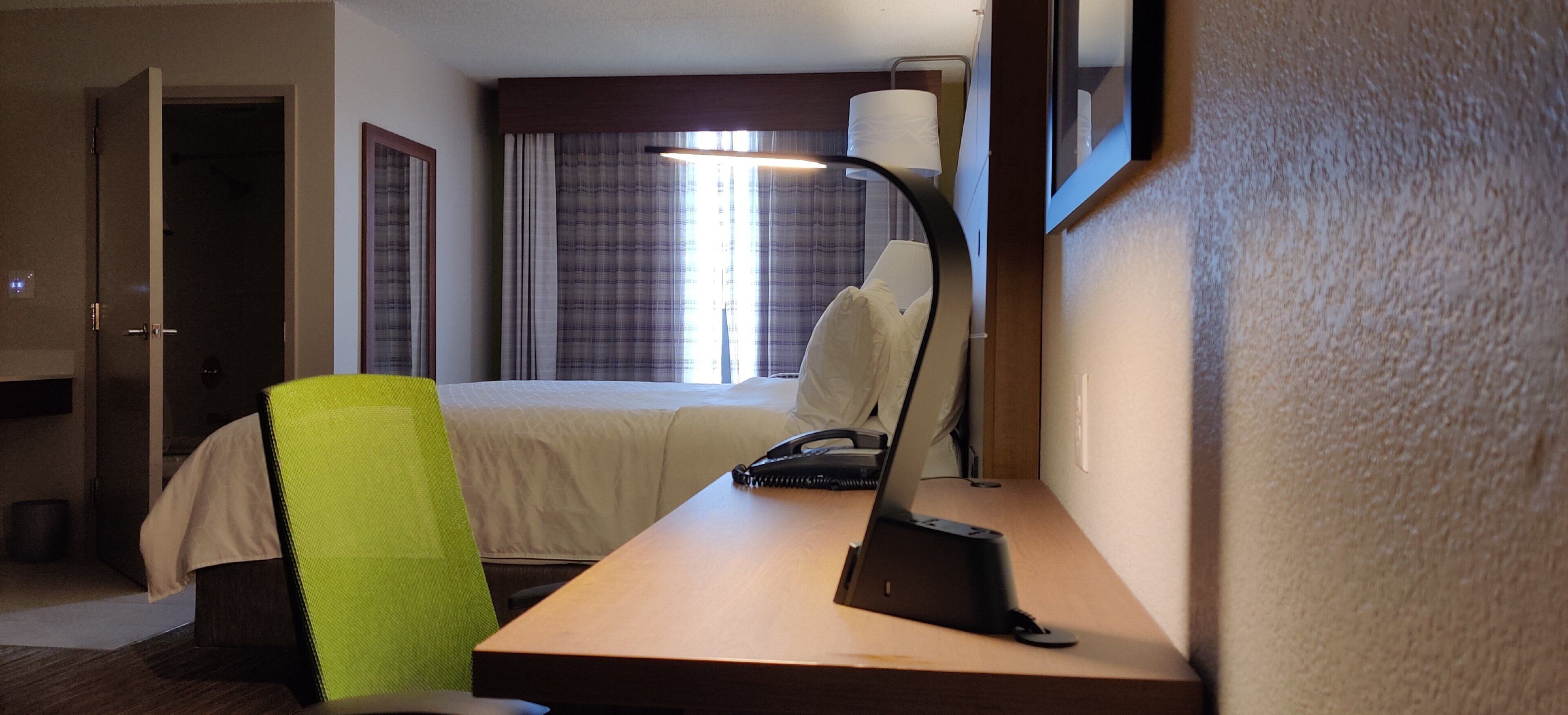 A hotel room with a bed, a desk, a chair, a lamp, and a window with curtains.