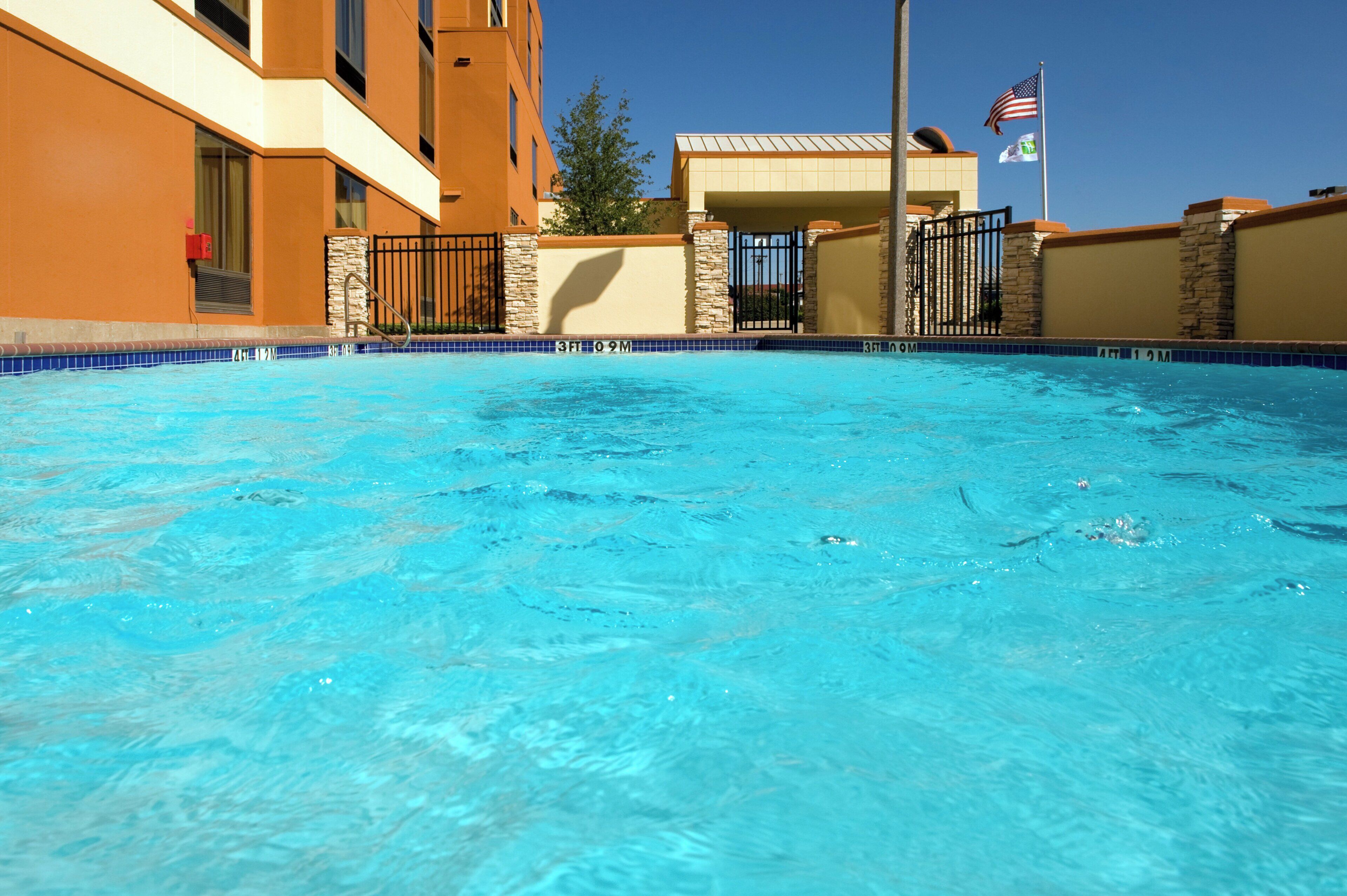 Una piscina con acqua cristallina e blu, circondata da un edificio con facciata in pietra e un’asta con la bandiera americana.