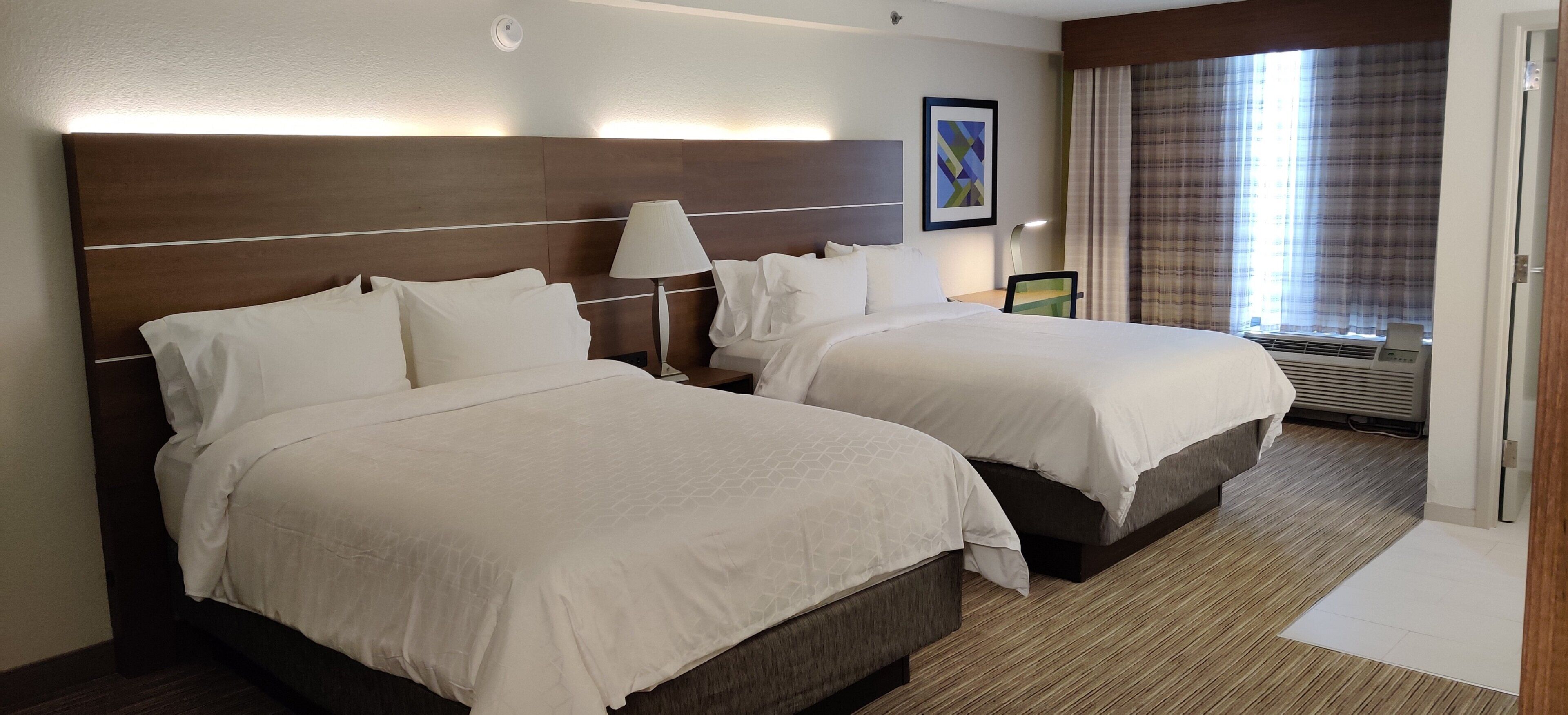A hotel room with two beds, a nightstand, a lamp, a window with curtains, and a wall-mounted picture.