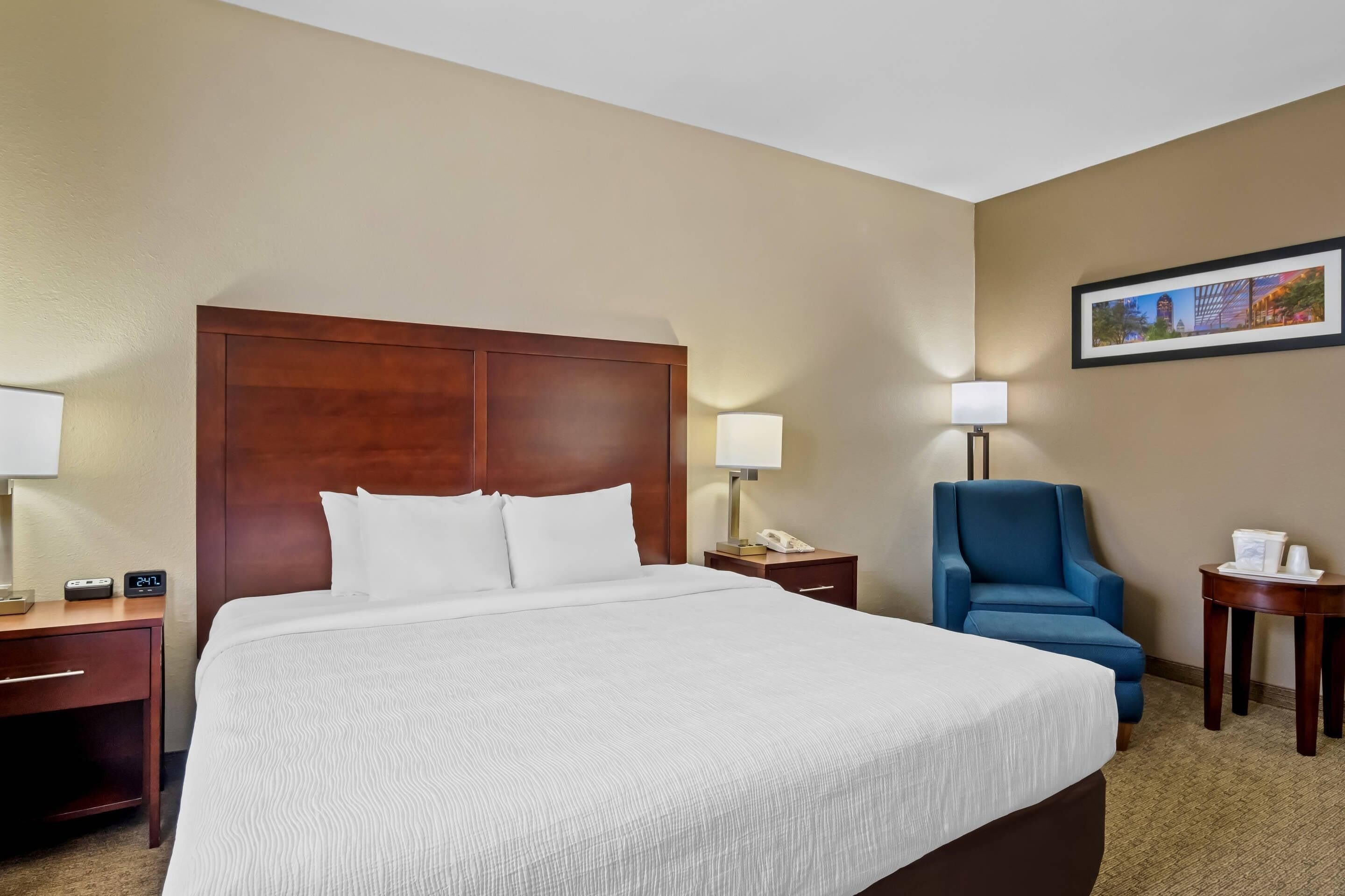 A hotel room with a large bed, a wooden headboard, a blue armchair, a small table with a lamp, and a framed picture on the wall.