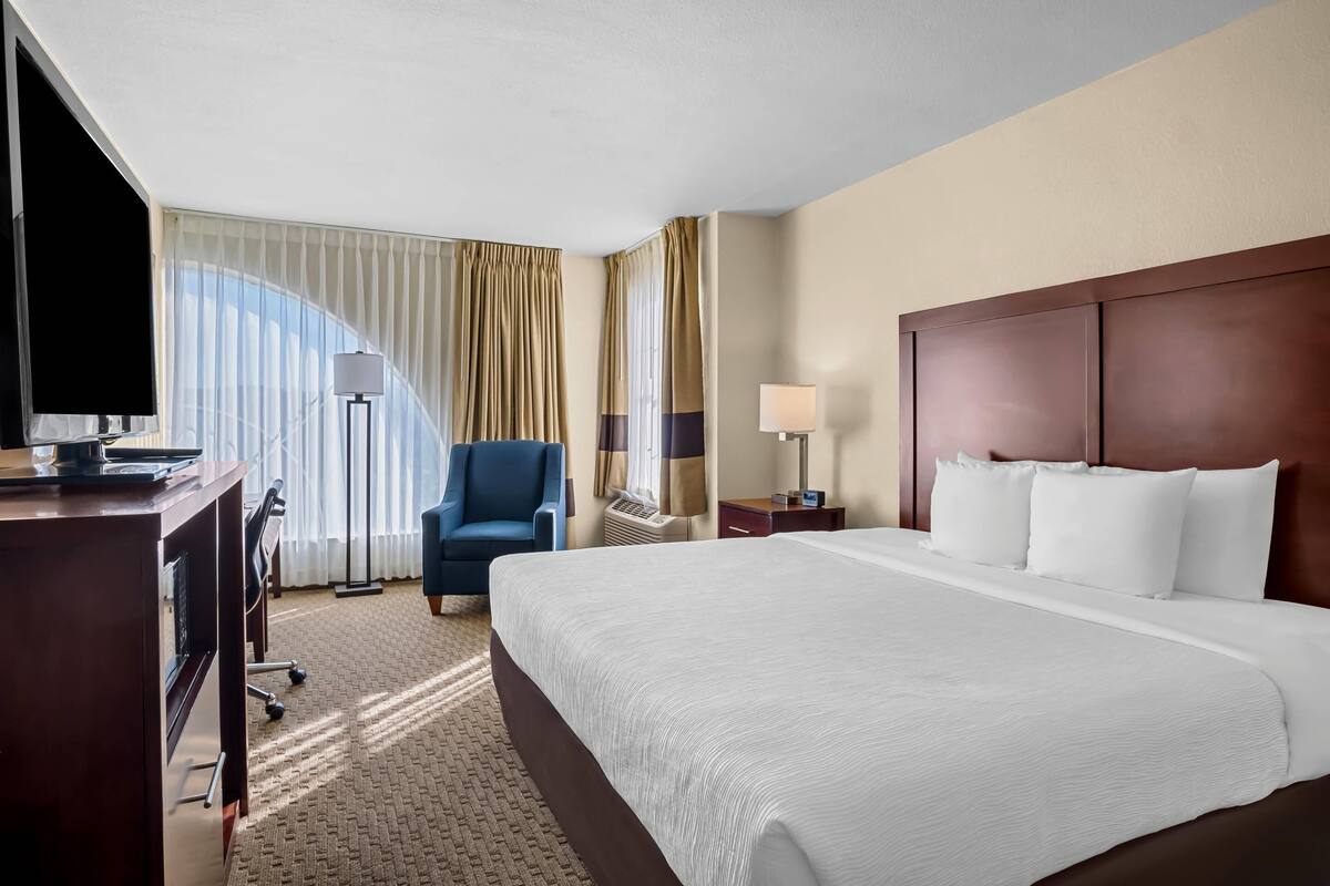 A hotel room with a large bed, a desk with a flat-screen TV, a chair, and a lamp.