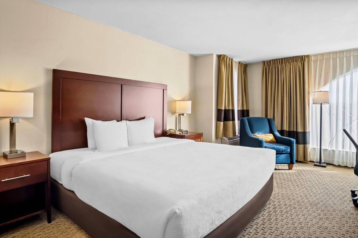A hotel room with a large bed, two bedside tables with lamps, a blue armchair, and a view of the outdoors through curtains.