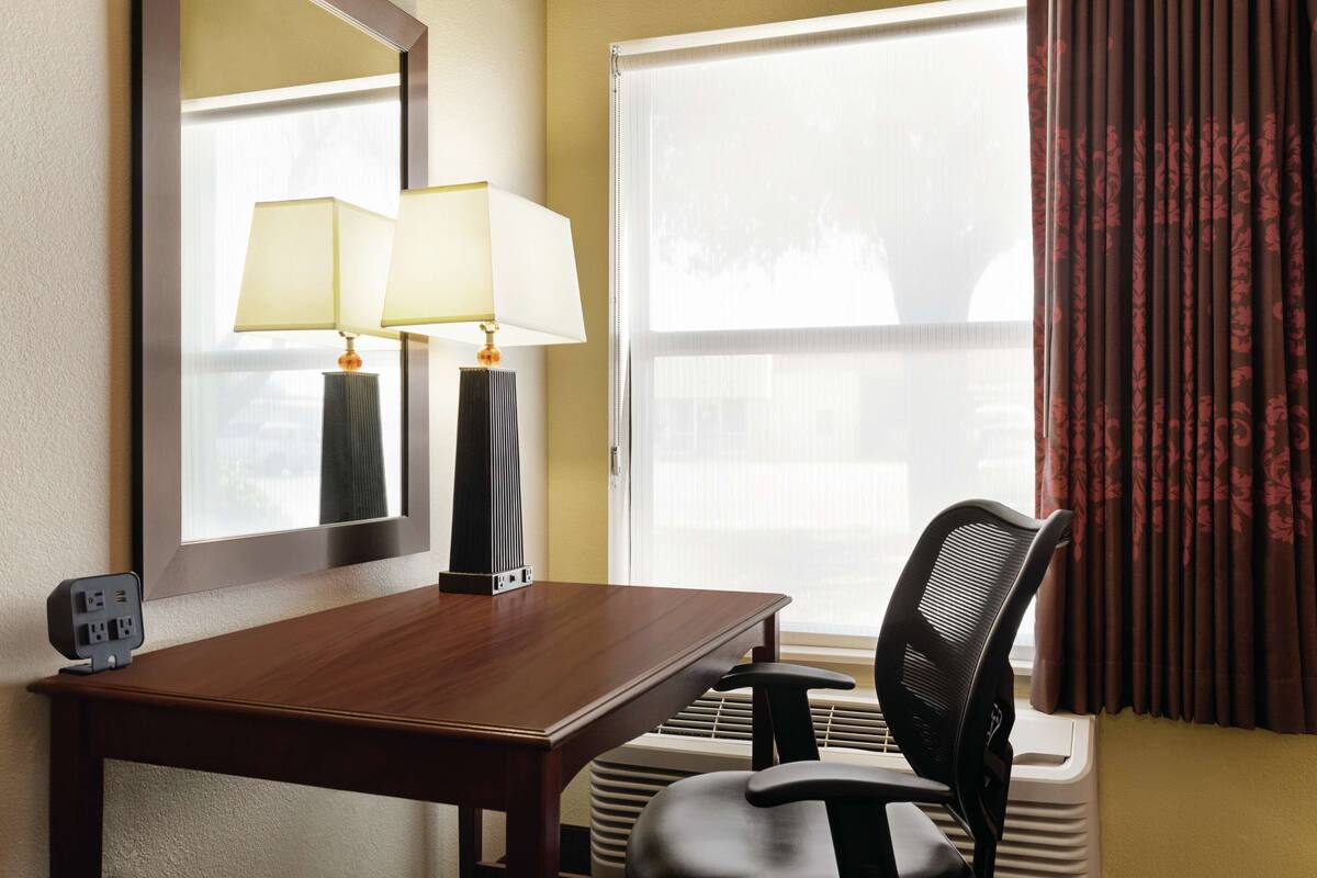 A hotel room with a wooden desk, two lamps, a chair, a large window with curtains, and a wall-mounted air conditioning unit.