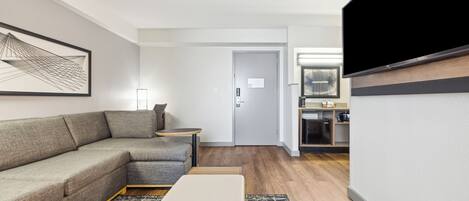 A modern hotel room with a sectional sofa, a small table, a white ottoman, a flat-screen TV mounted on the wall, and a kitchenette in the background.