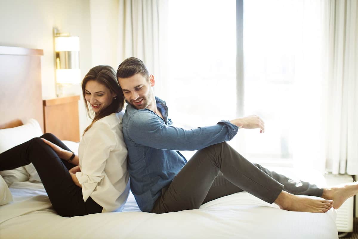 Un couple est assis sur un lit dans une chambre d’hôtel, près d’une table de chevet et d’une lampe.
