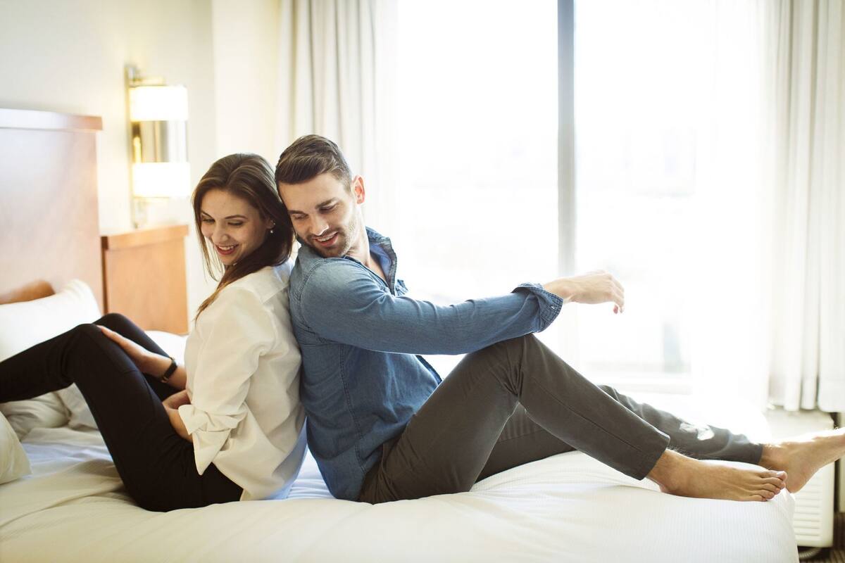 Un couple est assis sur un lit dans une chambre d’hôtel, près d’une table de chevet et d’une lampe.