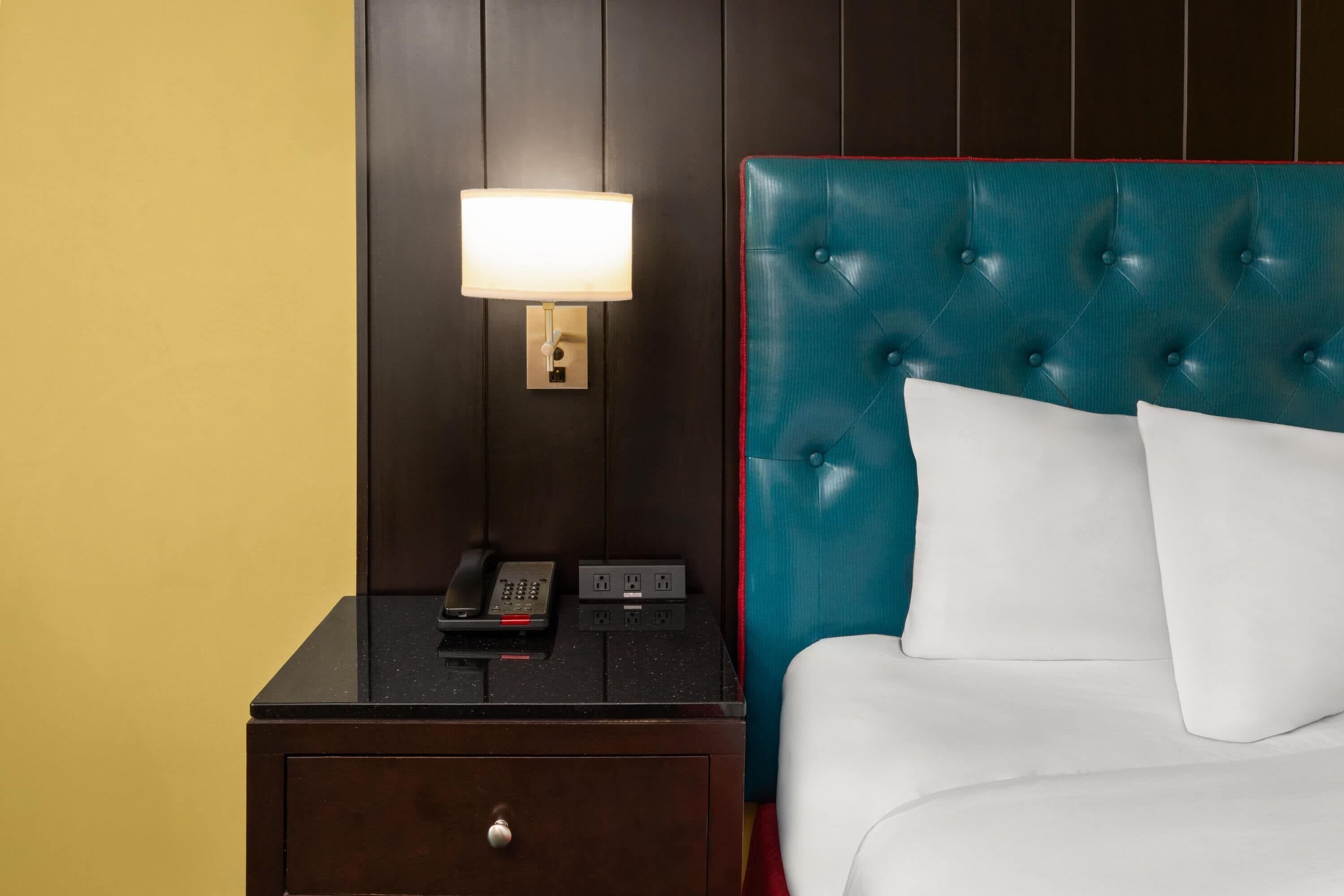 A hotel room with a bed, a nightstand with a lamp, a phone, and a drawer.