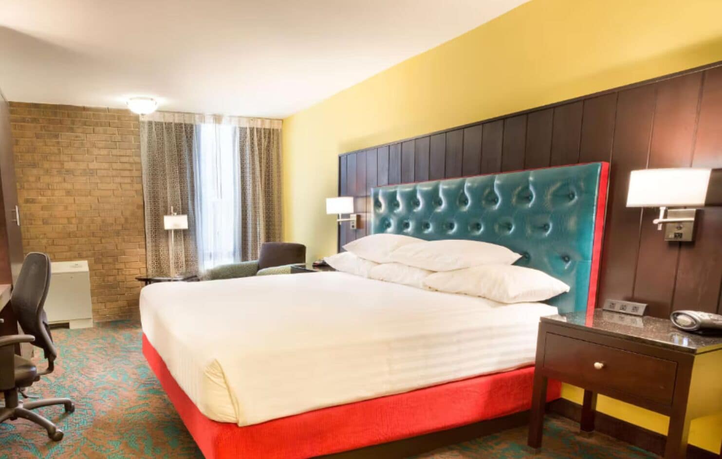 A hotel room with a large bed, a desk with a chair, a small table, and a wall-mounted lamp.