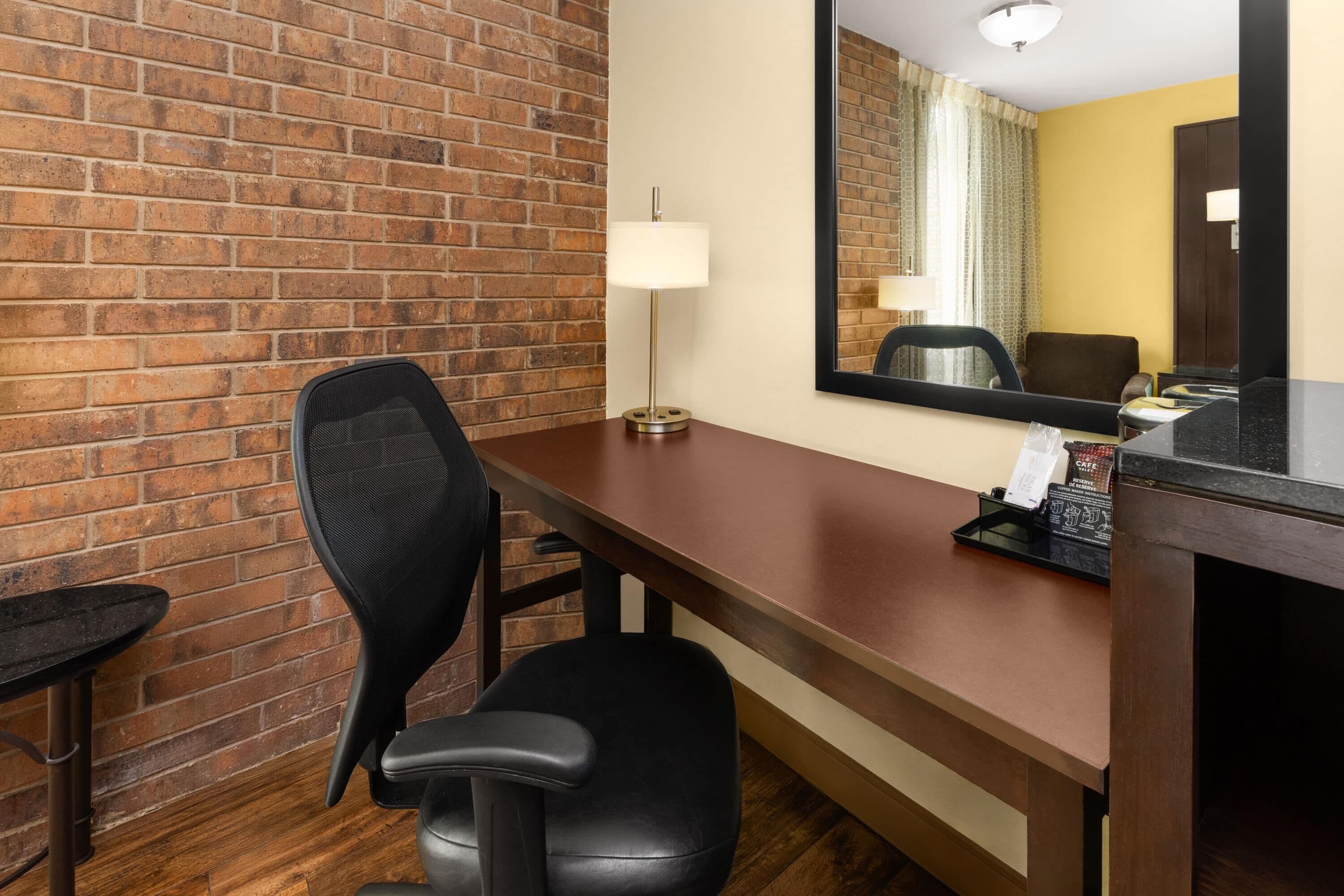 Une chambre d’hôtel avec un mur en briques, un bureau avec une lampe, une chaise, une petite table et un miroir.
