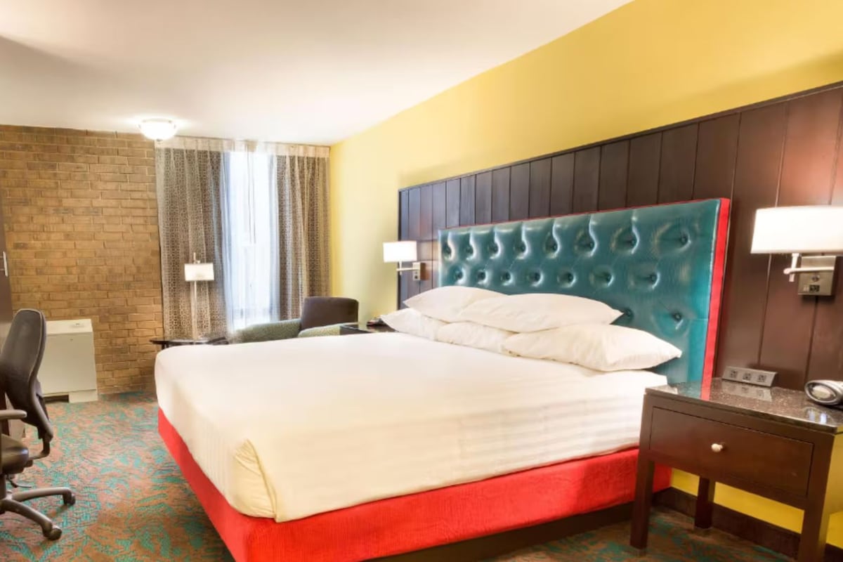 A hotel room with a large bed, a desk with a chair, a small table, and a wall-mounted lamp.