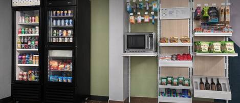 A self-service kiosk area with refrigerated beverage dispensers, a microwave, and shelves stocked with snacks, beverages, and packaged goods.