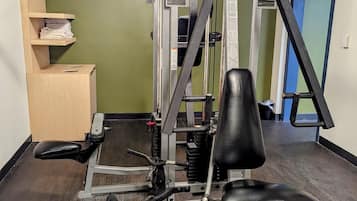 A gym with a squat rack, bench press, and weight stack. There is a wooden shelf with folded towels and a door in the background.