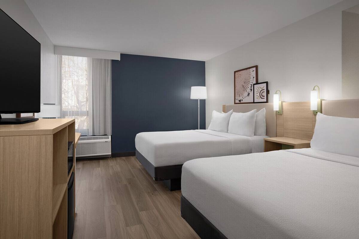 Ein Hotelzimmer mit zwei Betten, einem Fernseher, einem Schreibtisch und einem fensterbepflanzten Fenster.