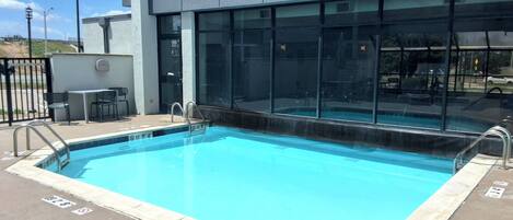 Una piscina rectangular al aire libre con agua azul cristalina, rodeada por una terraza de hormigón. Hay barandillas de metal en el borde de la piscina y una pared de vidrio con ventanas que reflejan el entorno.