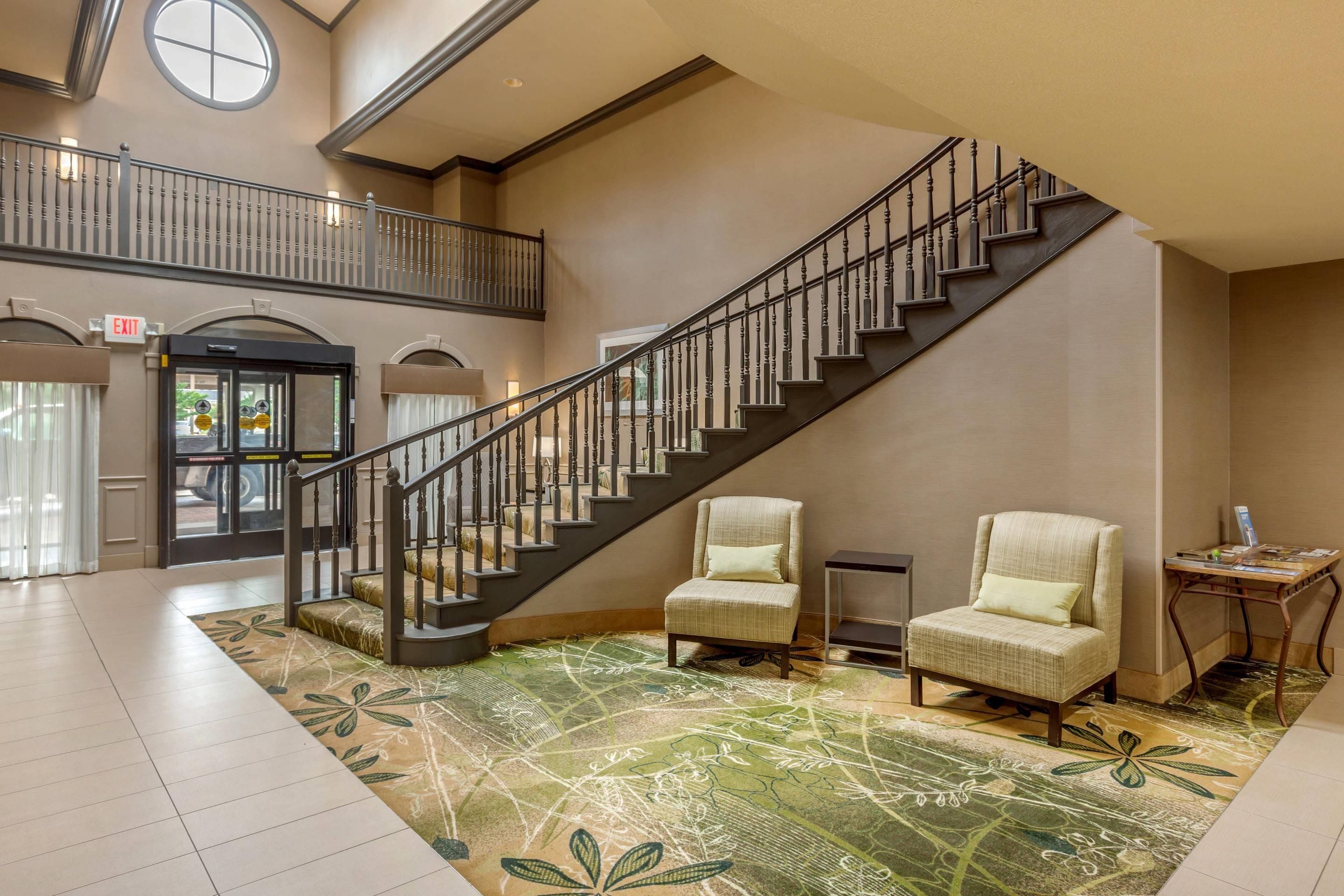 A spacious lobby with a central staircase, two armchairs, a small table, and a patterned carpet.